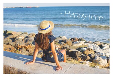 Beautiful girl in a hat sits near the sea on stones, the inscription of a happy time.の写真素材