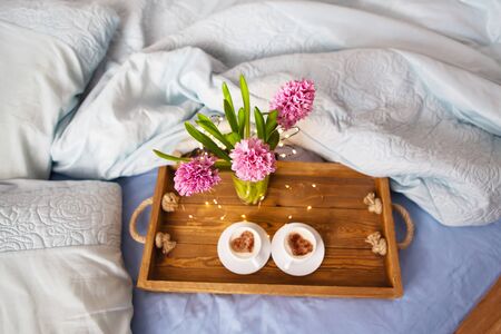 A bouquet of beautiful pink hyacinths is standing in a vase on a tray on which are two cups of cappuccino with a heart pattern, the concept of a surprise in bed, a beautiful sunny morning. View from above.の写真素材