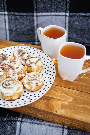 Two cups of black tea stand on a wooden tray on the sofa with a black and white checkered plaid. Fresh and fragrant cinnamon rolls close up lie on a plate with polka dots, beautiful morning.の写真素材