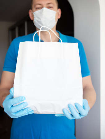 Home delivery service, masked and gloved guy delivers a bag of food to the door. Contactless delivery during a pandemic virus. Close-up.の写真素材