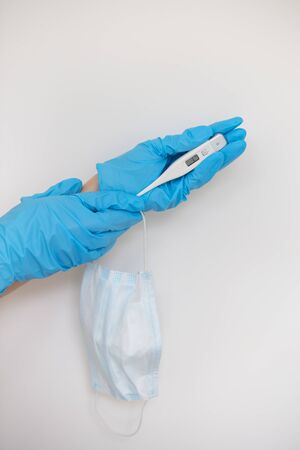 A medic's hand in a blue latex glove holds a thermometer and a medical mask showing the patient's...
