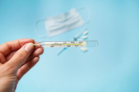 The hand holds a thermometer showing the patient's body temperature on a background of a medical mask, pills, syringe, on a blue background. Flu, illness conceptの写真素材