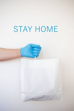 A hand in a blue rubber glove holds a grocery bag. Delivery man holding a paper bag in rubber gloves. Fast and safe delivery of goods to the door. The inscription stay home.の写真素材
