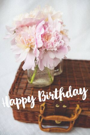 A very beautiful bouquet of pink peonies in a vase stands on a wooden suitcase. Beautiful composition. Inscription Happy Birthday.の写真素材