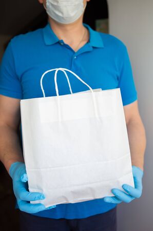 Delivery man holding a paper bag with groceries in rubber gloves and a protective mask. Fast and free delivery of goods to the door. Online store and express deliveryの写真素材