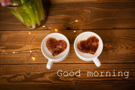 Two white cups of cappuccino stand on an old wooden tray. Garland, beautiful blinking. Top view, close-up. The inscription good morning.の写真素材