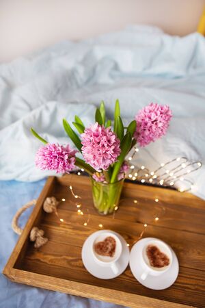 A bouquet of beautiful pink hyacinths is standing in a vase on a tray on which are two cups of cappuccino with a heart pattern, the concept of a surprise in bed, a beautiful sunny morning.の写真素材