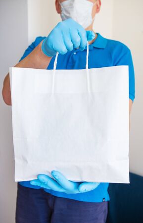 Home delivery service, masked and gloved guy delivers a bag of food to the door. Contactless delivery during a pandemic virus. Close-up.の写真素材