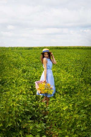 girl in a hat stands on a green field with a basket of flowers.の写真素材