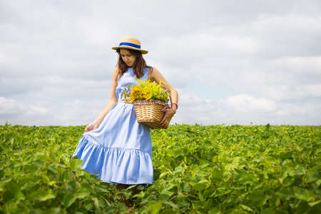 girl in a hat stands on a green field with a basket of flowers.の写真素材