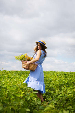 girl in a hat stands on a green field with a basket of flowers.の写真素材