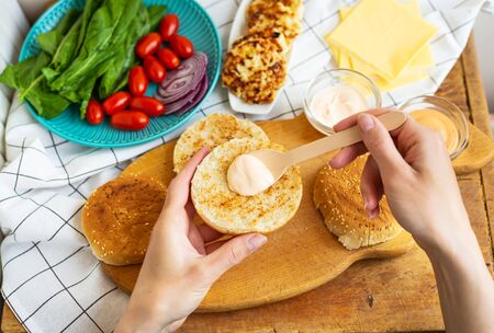 Preparation of all the ingredients for making a burger - bun, cutlet, cheese, salad, tomato, sauces. Top view, the girl s hands take a burger roll and spread the base with sauceの写真素材