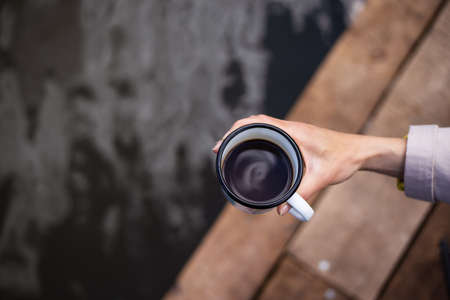 A girl holds a metal cup of coffee in her hands near the lake while sitting on a wooden pier. Close-up, top viewの写真素材