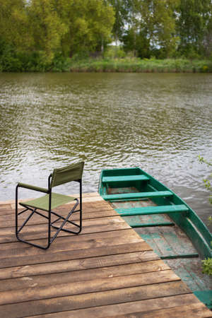 An empty chair and a fishing rod on a wooden pier by the lake, a green boat near the pier. Outdoor recreationの写真素材