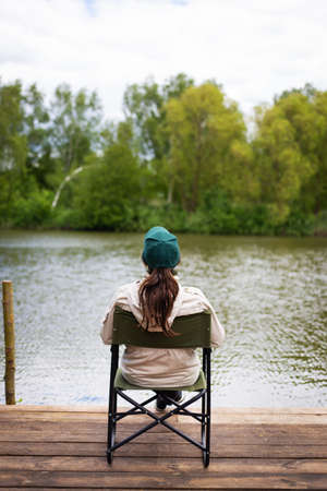 Girl sitting on the pier, fishing on the lake, smiling, rear view. Outdoor activities, beautiful summer dayの写真素材