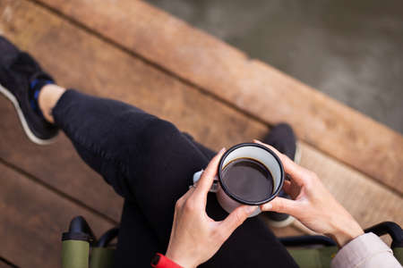 A girl holds a metal cup of coffee in her hands near the lake while sitting on a wooden pier. Close-up, top viewの写真素材
