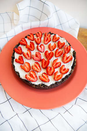 Strawberry cheesecake decorated with heart-shaped strawberries lies on a coral-colored plate, stands on a napkin and a wooden table. Illustration of a cheesecake recipe with strawberriesの写真素材