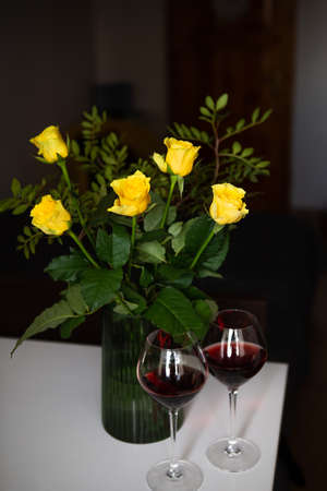 A very beautiful bouquet of yellow roses stands in a green vase and two glasses of red wine. Romantic eveningの写真素材
