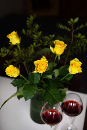 A very beautiful bouquet of yellow roses stands in a green vase and two glasses of red wine. Romantic eveningの写真素材