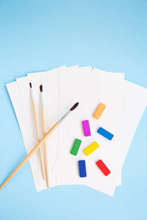 Multi-colored paints, watercolor brushes of different sizes lie together with watercolor paper on a blue background. Drawing lessons. View from aboveの写真素材