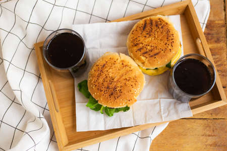Big and tasty burgers lie on a wooden tray with a glass of cola. Wooden table and white checkered napkin. View from aboveの写真素材