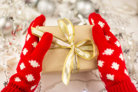 Female hands hold a Christmas gift with a gold ribbon in red mittens, new year mood, preparation for the celebrationの写真素材