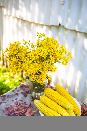 Boiled corn lies on a plate on the table along with a bouquet of yellow flowers. Sunny summer dayの写真素材