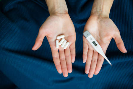 The girl is holding an electronic thermometer and pills for the treatment of the new COVID-19 virus. Stay home, save your life. Place for an inscription. Close-up.の写真素材