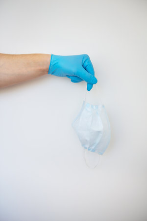 A hand in blue gloves holds a white medical protective mask on a white background. Protection against viruses and flu. Close-up. Place for an inscription.の写真素材