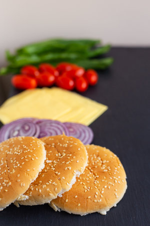 Preparation for the preparation of a hamburger, all ingredients: cheese, salad, tomato, onion, bun lie on a black stone background. Close-upの写真素材
