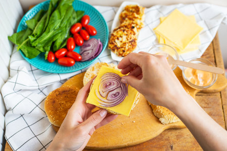 Preparation of all the ingredients for making a burger - bun, cutlet, cheese, salad, tomato, sauces. Top view, girl's hands take a burger bun and lay on a purple bowの写真素材
