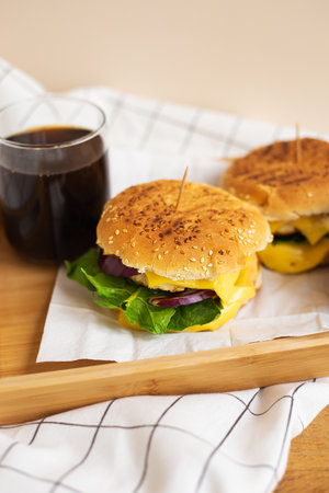Large and tasty grilled hamburgers with sesame seeds lie on a wooden tray with a glass of cola. Wooden table and white checkered napkin. Close-upの写真素材