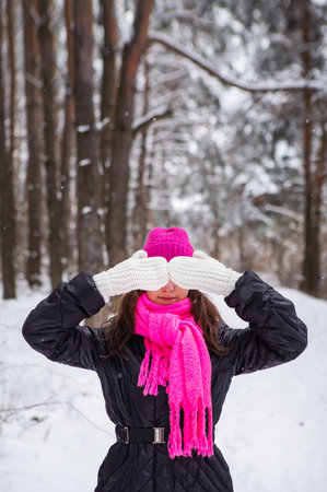 A girl in a pink scarf and hat stands in the middle of a pine forest and covers her face with white mittens. Beautiful pine forest on a frosty dayの写真素材