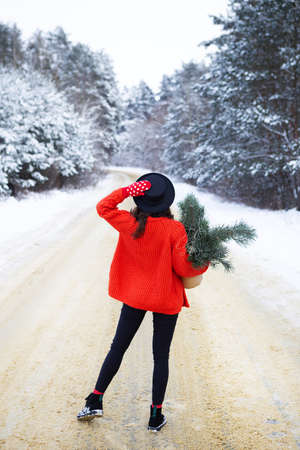 A girl in a red sweater and hat stands in the middle of a snowy road in a forest with pine branches. Weekend tripの写真素材