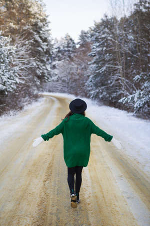 A young girl in a green sweater and hat walks in the middle of a snowy road in a thick pine forest. Freezing dayの写真素材