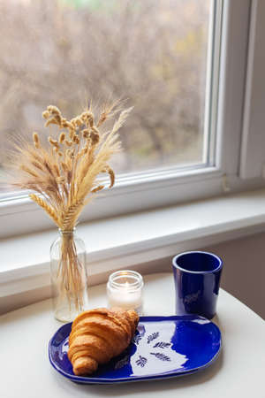 Handmade blue clay plate with croissant on it. Vase with wheat and candles