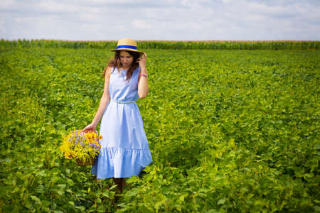 girl in a hat walks on a green field with a basket of flowers.の写真素材