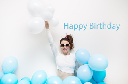 Happy girl in glasses smiles and holds blue balloons in her hands. Place for an inscription, holiday concept. Happy birthday letteringの写真素材