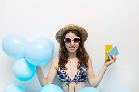 Happy girl in a striped swimsuit, hat holds a passport and balloons for a trip to the sea against the background of blue balloons. Place for an inscription, vacation conceptの写真素材