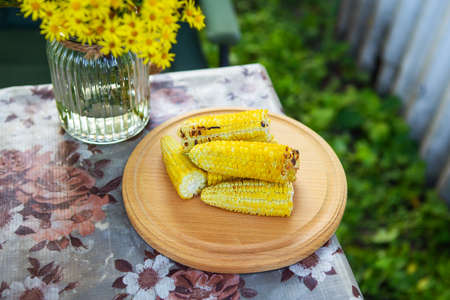 Delicious, sweet young grilled fried corn lies on a wooden board on the table along with a bouquet of flowers, outdoor recreationの写真素材