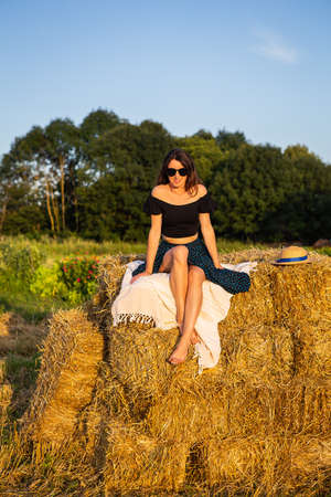 Young beautiful woman in black glasses looks at the setting sun, sits on a large pile of straw balesの写真素材