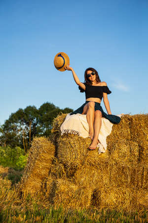 Young beautiful woman in black glasses looks at the setting sun and holds a straw hat, sits on a large pile of straw balesの写真素材