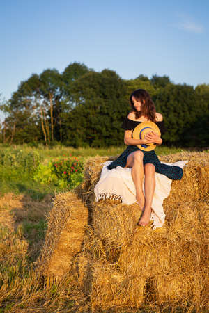Young beautiful woman looks at the setting sun and holds a straw hat, sits on a large pile of straw balesの写真素材