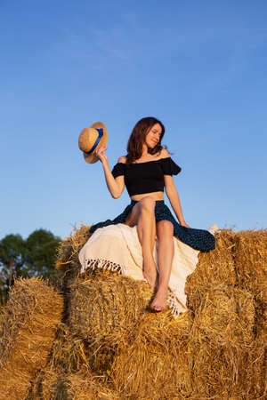 Young beautiful woman looks at the setting sun and holds a straw hat, sits on a large pile of straw balesの写真素材