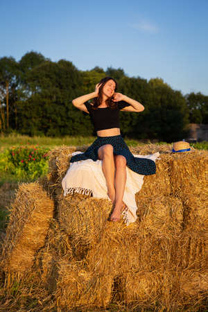 Young beautiful woman looks at the setting sun and holds a straw hat, sits on a large pile of straw balesの写真素材