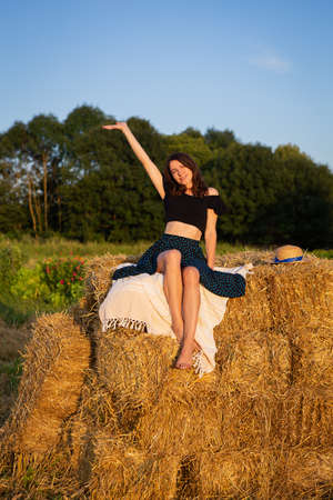 Young beautiful woman in a straw hat looks at the setting sun, the girl sits on a large pile of straw bales. Warm summer eveningの写真素材