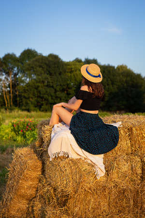 A young beautiful woman in a straw hat stands on a large pile of straw bales. Freedom, wind, setting sunの写真素材