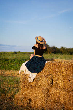 Young beautiful woman in a straw hat looks at the setting sun, the girl sits on a large pile of straw bales. Warm summer eveningの写真素材