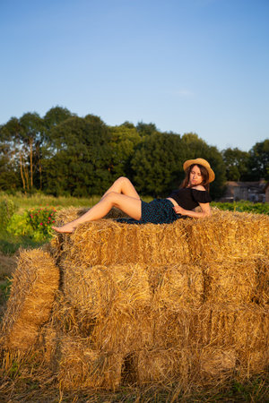 Young beautiful woman in a straw hat looks at the setting sun, the girl sits on a large pile of straw bales. Warm summer eveningの写真素材