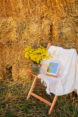 Picnic in the field near straw bales. The setting sun. Rustic style - wood chair, plaid, bouquet of flowers, mirror. Romantic dateの写真素材